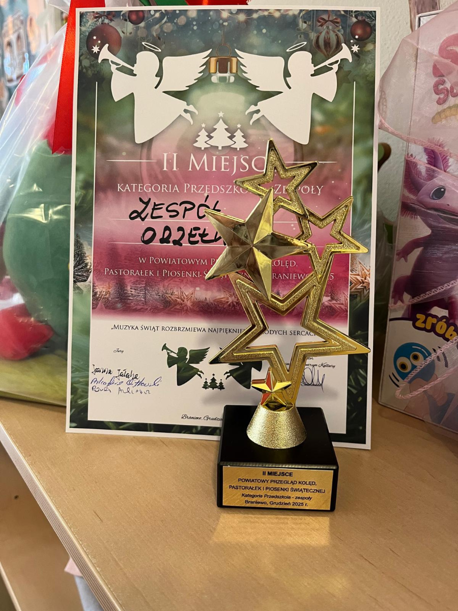 Złota statuetka w kształcie gwiazd stoi na drewnianym blacie. Przed nią oparta jest pamiątkowa plansza z informacją o II miejscu w przeglądzie kolęd i pastorałek w kategorii przedszkola zespoły