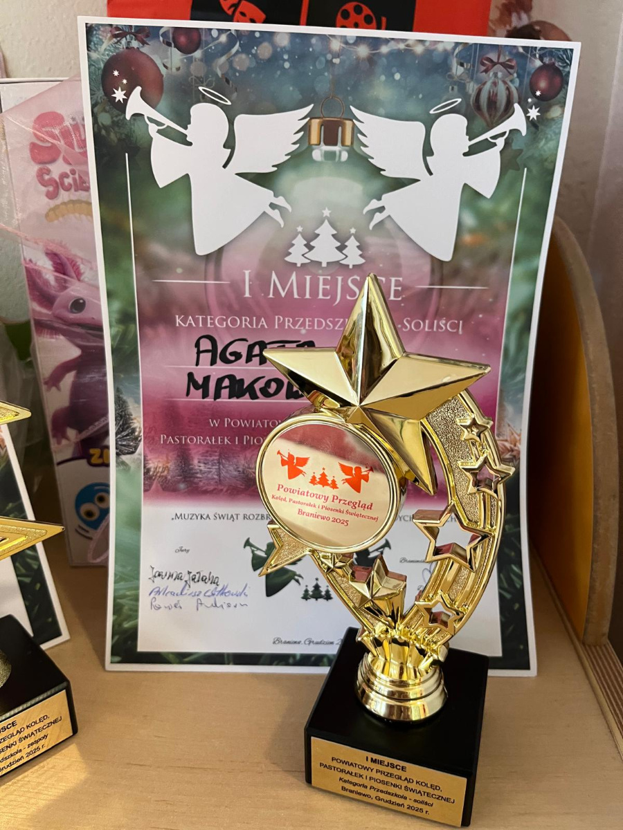 Złota statuetka z motywem gwiazdy stoi na drewnianym blacie. Za nią widoczny jest dyplom informujący o I miejscu w Powiatowym Przeglądzie Kolęd w kategorii przedszkola sol