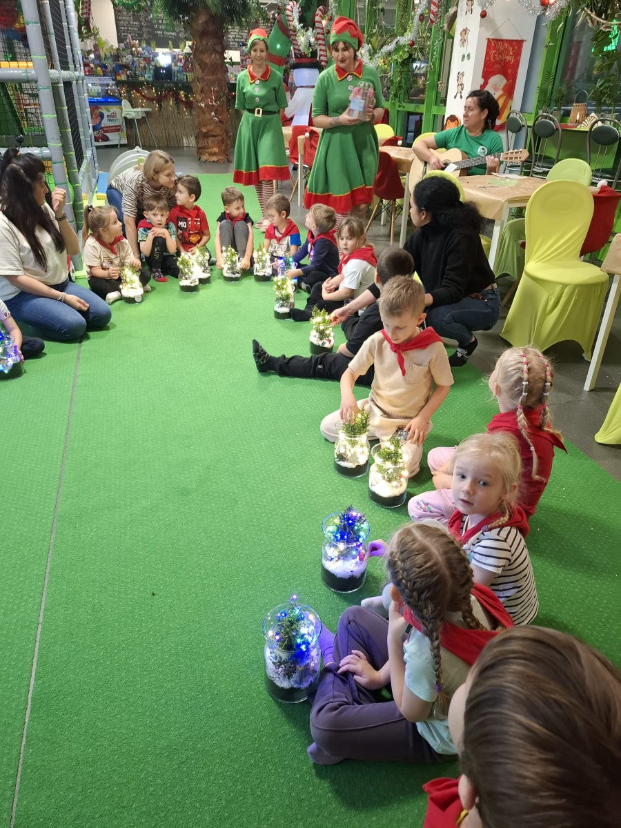 Dzieci siedzą w kole na zielonej macie przed nimi świecą lampiony z małymi choinkami. Obok dorośli w tle elfy w zielonych strojach