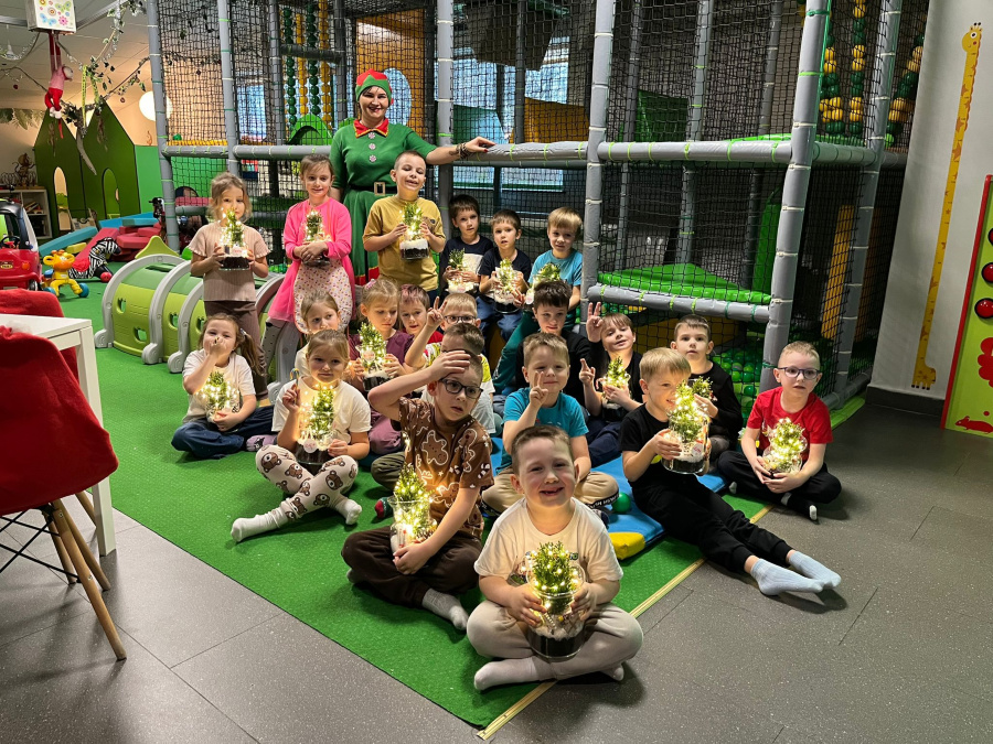 Grupa dzieci siedzi i stoi na zielonej macie, trzymając lampiony z małymi, świecącymi choinkami. Za nimi stoi dorosła osoba przebrana za elfa. Dzieci uśmiechają się do aparatu
