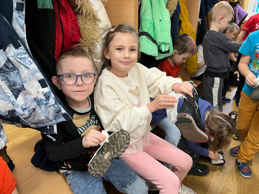 Dwoje dzieci siedzi obok siebie na niskiej ławce w szatni. Trzymają w dłoniach buty i wycierają je chusteczkami