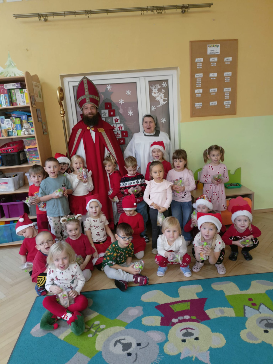 Grupa dzieci pozuje do wspólnego zdjęcia. Część dzieci stoi część siedzi na dywanie mają na sobie ubrania w świątecznych kolorach. Za dziećmi stoi mężczyzna w czerwonym 
