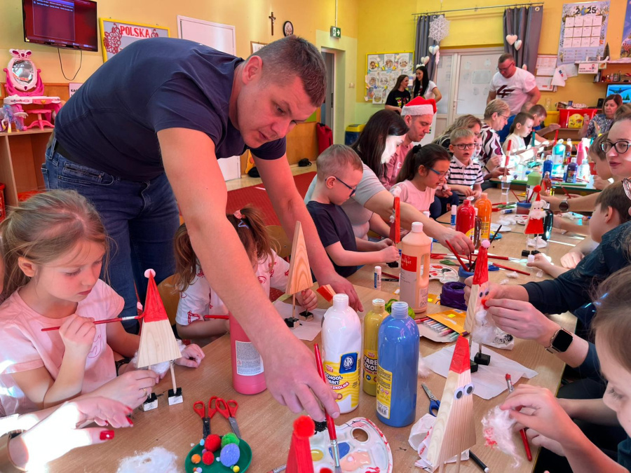 Przy długim stole w sali dzieci siedzą obok dorosłych i wspólnie malują drewniane figurki. Dzieci trzymają pędzle nanoszą farbę i doklejają elementy dekoracyjne