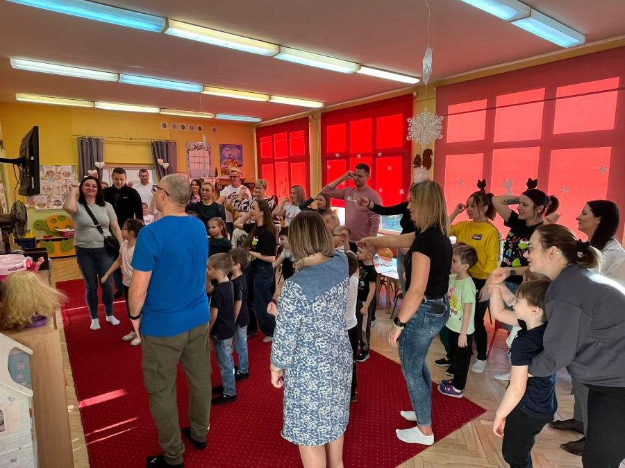 W sali dzieci i dorośli stoją razem na dywanie ustawieni w luźnej grupie. Wszyscy wykonują te same ruchy rąk i ciała naśladując prowadzącego. Dzieci obserwują dorosłych i aktywnie uczestniczą w zabaw