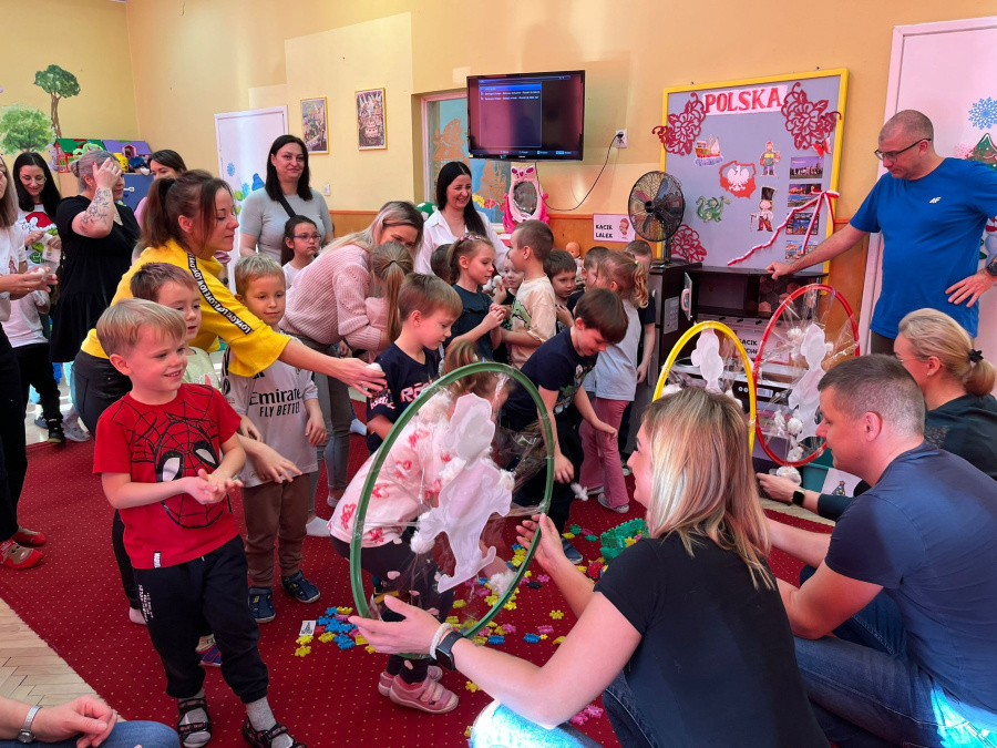 W sali dzieci stoją w półkolu i podchodzą kolejno do obręczy trzymanych przez dorosłych. W obręczach zawieszone są lekkie, białe materiały