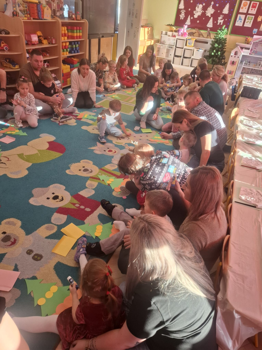 Dzieci i dorośli siedzą w dużym kręgu na dywanie w sali przedszkolnej. Dzieci przyklejają papierowe elementy tworząc choinki a dorośli siedzą obok i pomagają w pracy. Na podłodze leżą kartki kleje 