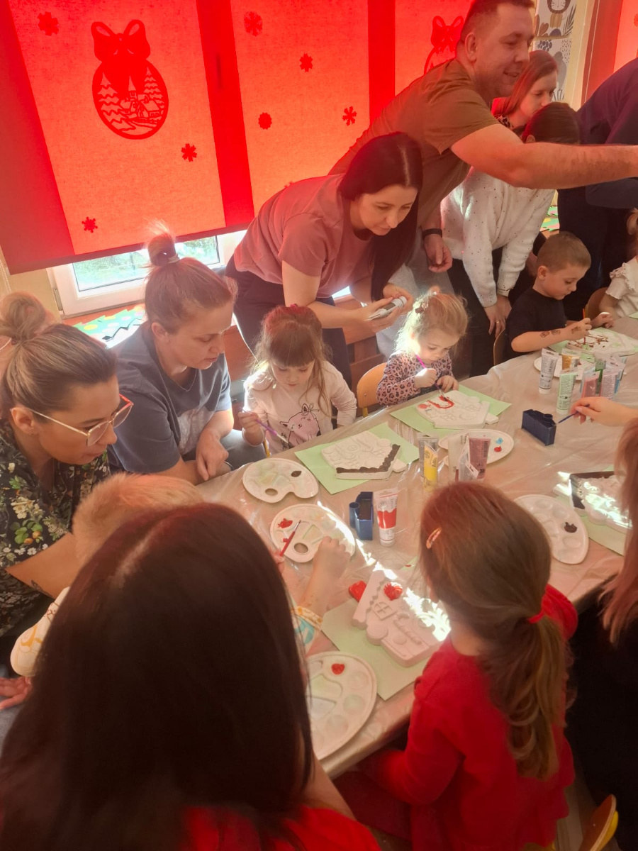 Dzieci siedzą przy długim stole i malują gipsowe ozdoby farbami. Obok nich stoją dorośli którzy pomagają i podają materiały. Na stole leżą palety pędzle i tubki z farbami