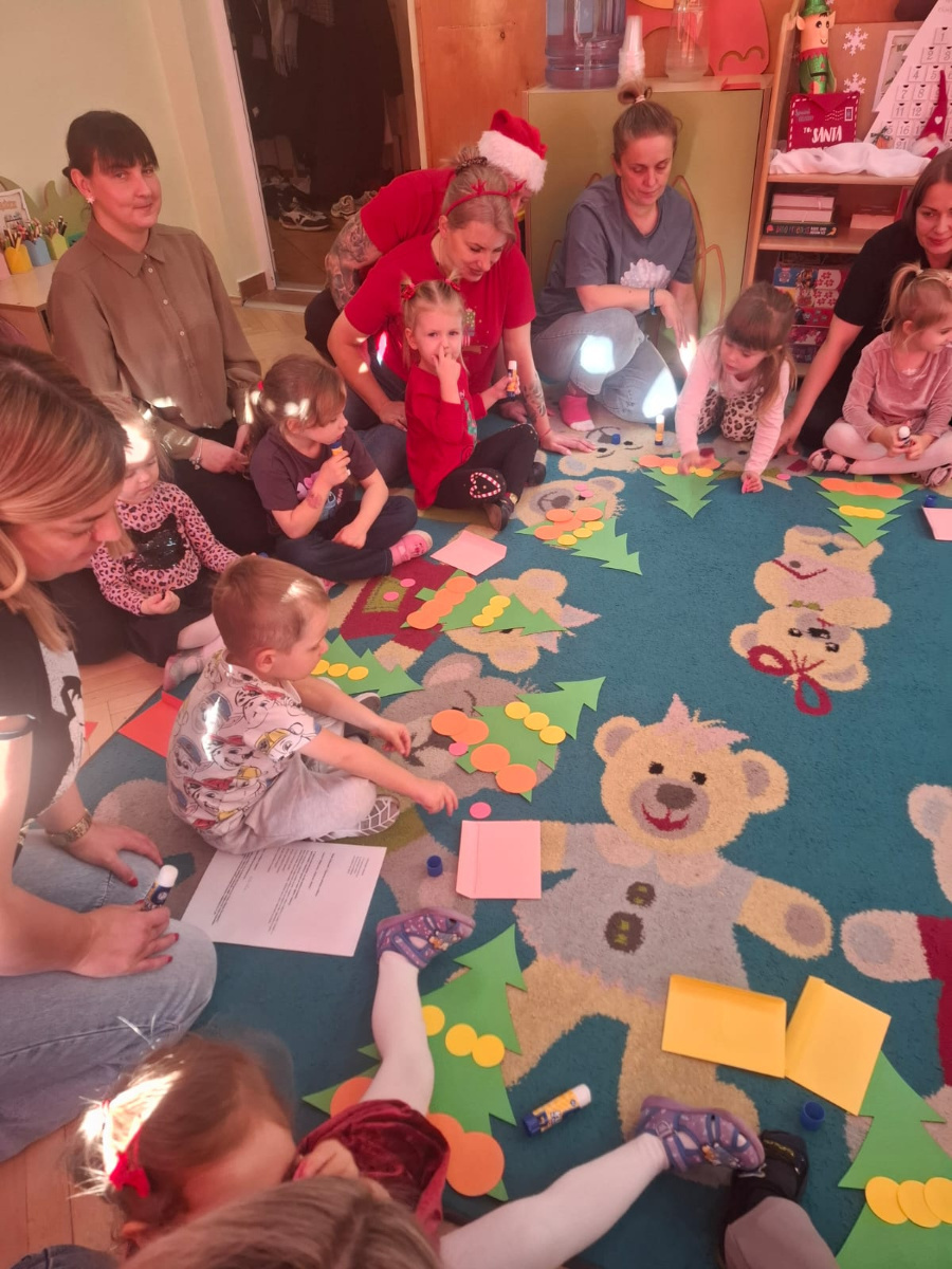 Dzieci siedzą w kręgu na dywanie i przyklejają papierowe elementy, tworząc choinki. Wokół nich znajdują się kartki, kleje i kolorowe kółka. Obok siedzą dorośli, którzy obserwują i wspierają dzieci