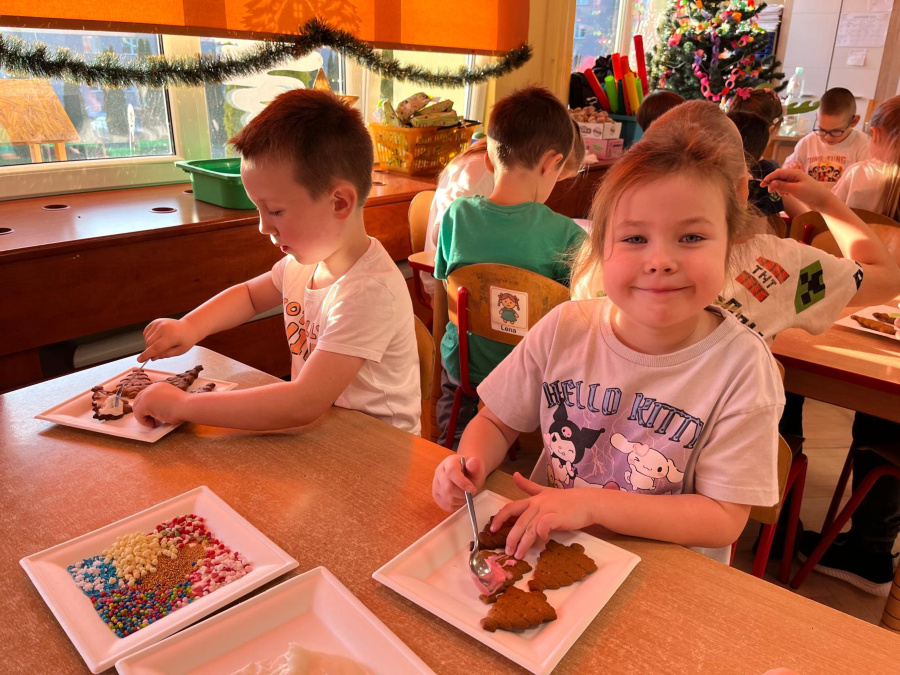 Dzieci siedzą przy stolikach i dekorują pierniki kolorowymi posypkami oraz lukrem. Skupione twarze małe dłonie pracują z łyżeczkami. W tle świąteczne ozdoby i choinka
