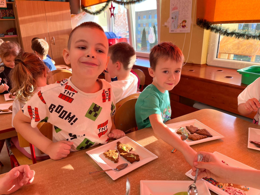 Dzieci siedzą przy stolikach i dekorują pierniki lukrem oraz posypkami. Jedno dziecko uśmiecha się i spogląda w stronę aparatu inne skupione jest na pracy przy talerzyku