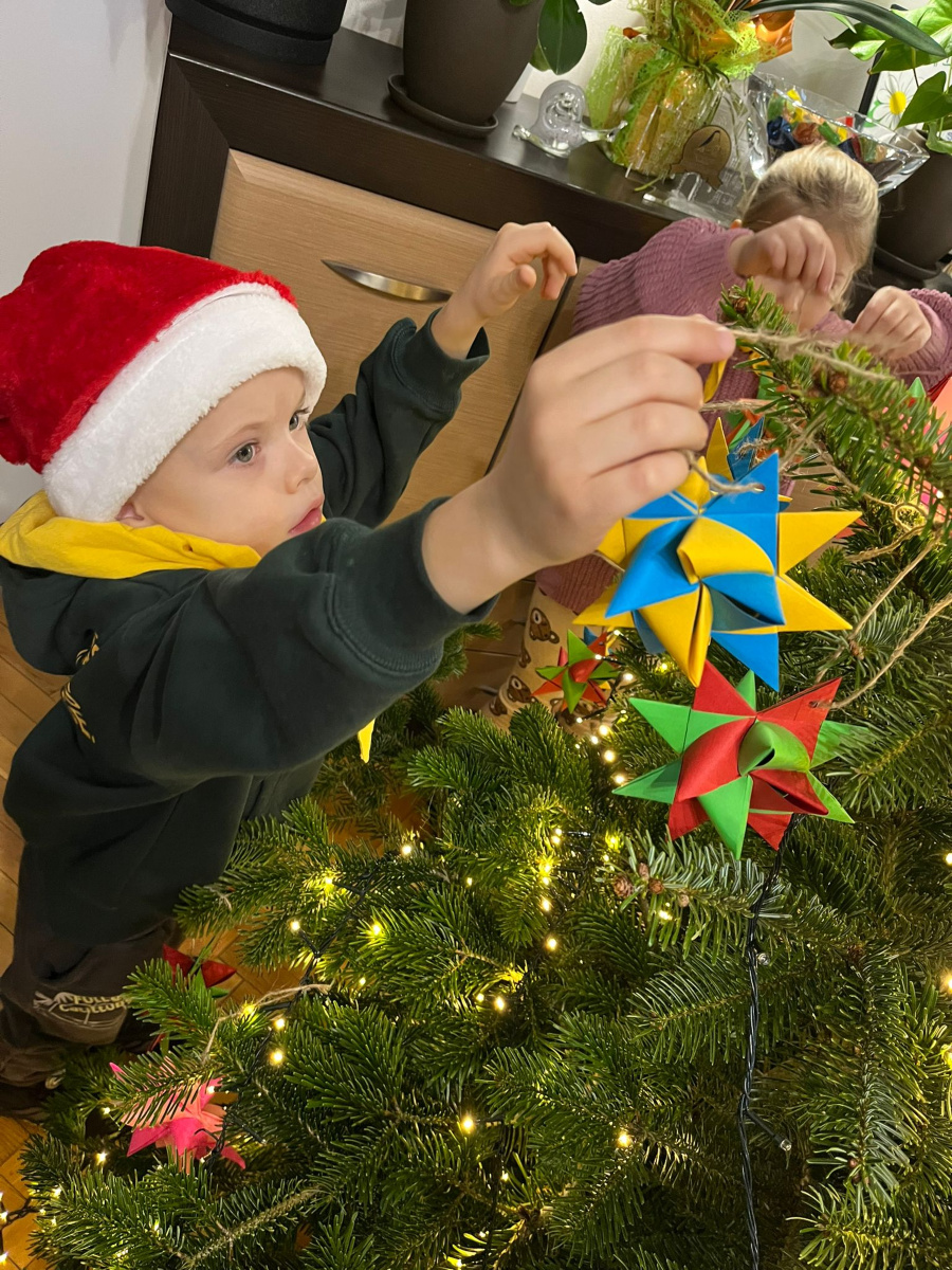 Dwoje dzieci stoi przy choince ustawionej w pomieszczeniu. Jedno z dzieci w czerwonej czapce i żółtym szaliku sięga rękami w stronę gałązek zawieszając kolorową papierową ozdobę