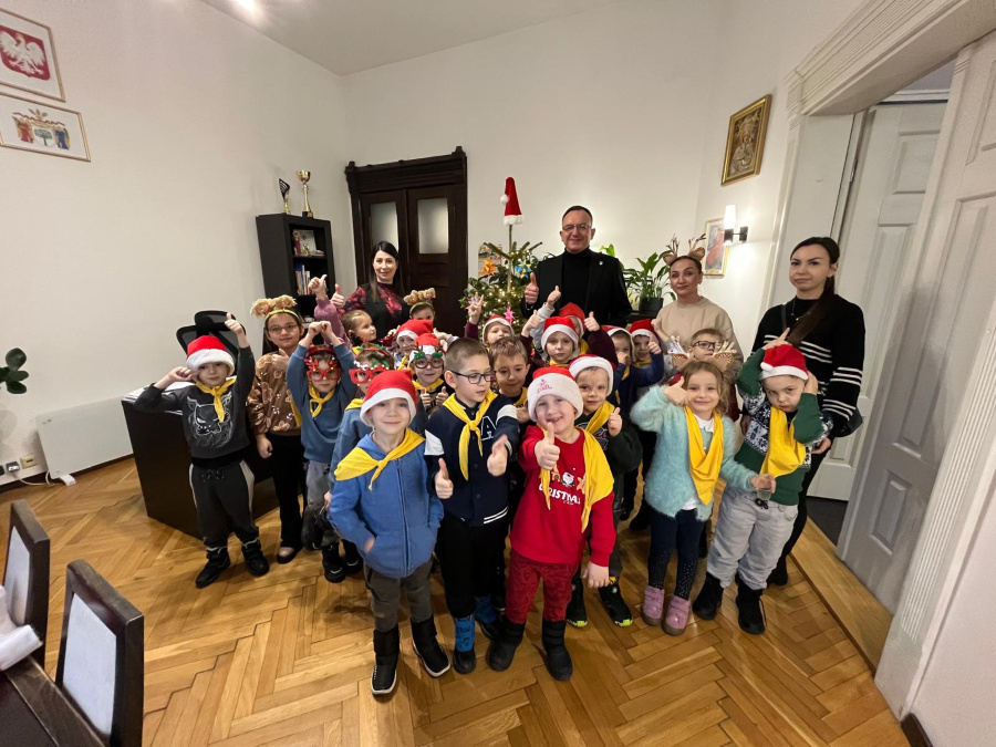 Grupa dzieci stoi blisko siebie w pomieszczeniu z parkietową podłogą. Wiele dzieci ma na głowach czerwone czapki, część trzyma uniesione kciuki w górę 