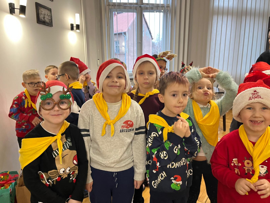 Grupa dzieci stoi w pomieszczeniu ustawiona blisko siebie. Kilkoro dzieci ma na głowach czerwone czapki część nosi żółte chusty na szyi niektóre mają świąteczne okulary lub opaski