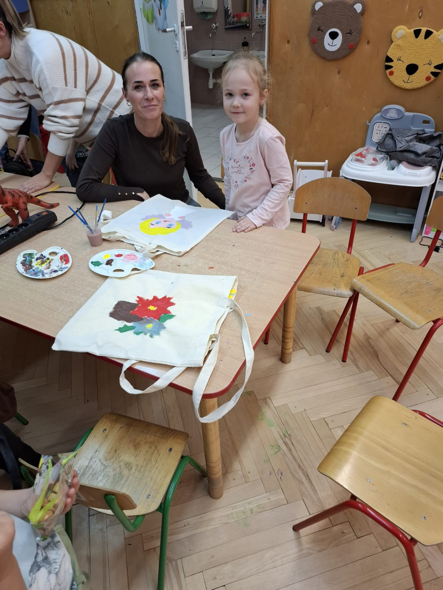 Przy stoliku w sali przedszkolnej dziecko stoi obok dorosłej osoby. Razem prezentują wykonane prace plastyczne namalowane farbami na materiałowych torbach. Na blacie leżą palety z farbami i pędzle