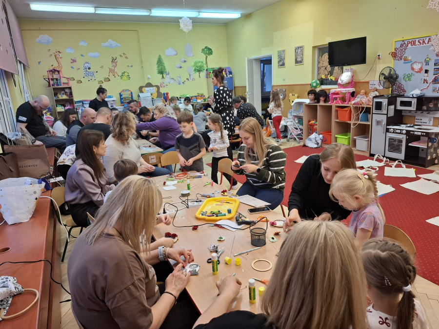 W sali dzieci siedzą i stoją przy stolikach razem z dorosłymi. Wspólnie wykonują prace plastyczne wycinają kleją nawlekają elementy i dekorują drobne przedmioty. W tle widać zabawki