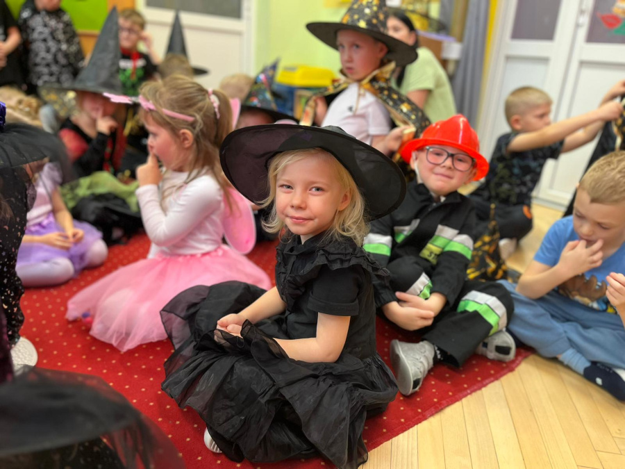 Kilka dzieci siedzi na dywanie w sali. Na pierwszym planie dziecko siedzi po turecku ubrane na czarno ma na głowie kapelusz i patrzy w stronę aparatu