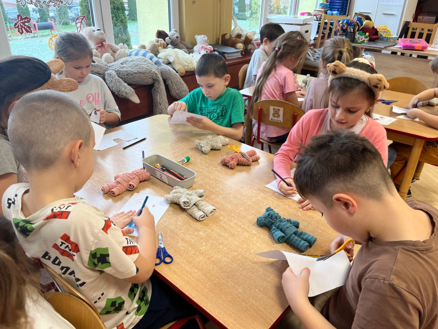 Kilka dzieci siedzi przy stolikach i pracuje trzymają kartki papieru nożyczki i kredki wycinają oraz rysują. Na blatach leżą zwinięte kawałki materiału