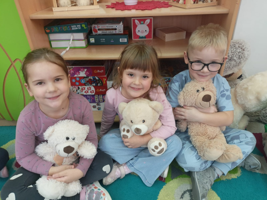 Troje dzieci siedzi obok siebie na dywanie. Dzieci trzymają w ramionach pluszowe misie i patrzą w stronę aparatu