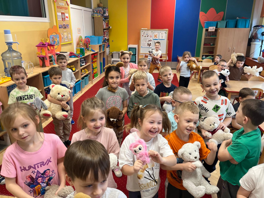 W sali stoi duża grupa dzieci. Dzieci trzymają w rękach pluszowe misie i inne maskotki. Część dzieci stoi blisko siebie inne są lekko rozproszone