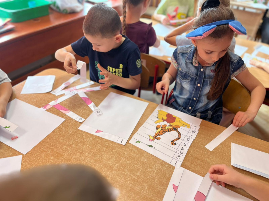 Dwoje dzieci siedzi przy stoliku i układa obrazki pocięte na paski z numerami. Dziewczynka w opasce z niebieskimi uszami składa ilustrację z Tygryskiem z Kubusia Puchatka
