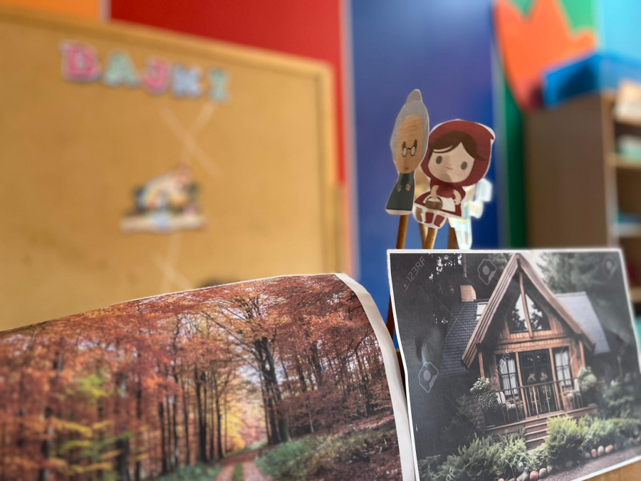 Na pierwszym planie znajdują się ilustracje przedstawiające las i drewniany domek oraz papierowe postacie Czerwonego Kapturka i babci umieszczone na patyczkach