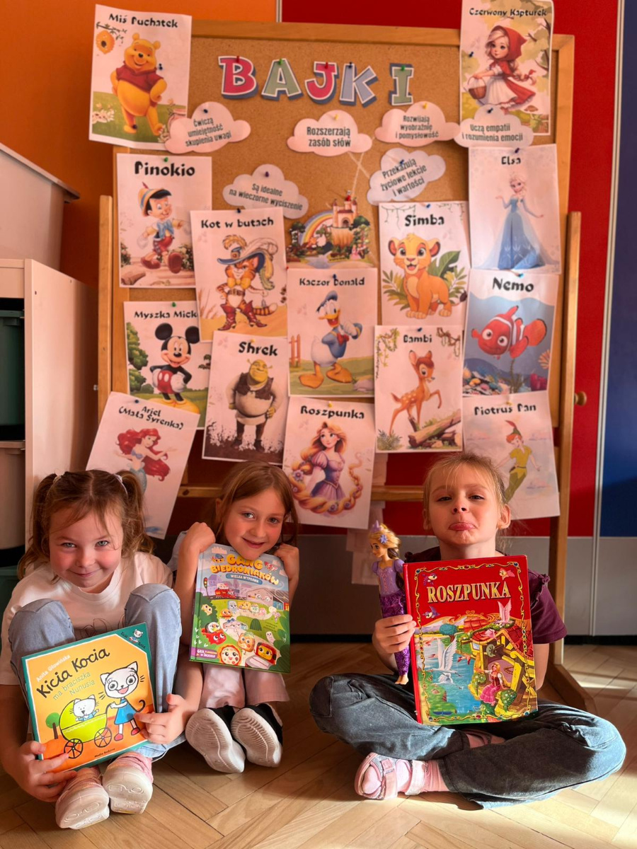 Trzy dziewczynki siedzą na podłodze przed tablicą z napisem BAJKI. Na tablicy znajdują się ilustracje i napisy z tytułami znanych bajek takich jak Miś Puchatek Pinokio