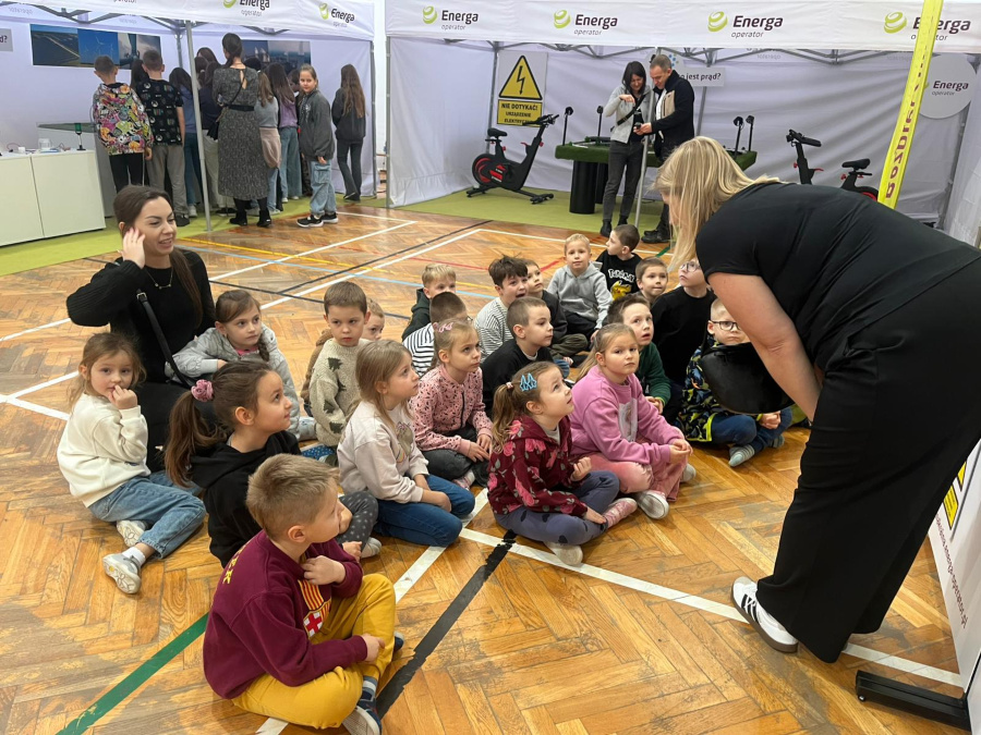 Grupa dzieci siedzi na podłodze w półkolu. Dzieci patrzą w jednym kierunku