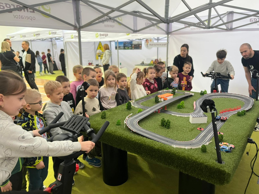 Grupa dzieci stoi blisko siebie wokół dużego stołu z makietą toru wyścigowego. Dzieci patrzą przed siebie, część z nich pochyla się z zaciekawieniem, niektóre wskazują elementy makiety