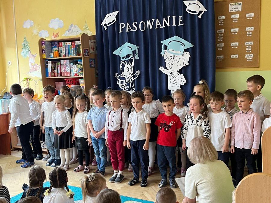 Dzieci stoją w rzędzie i śpiewają podczas uroczystości pasowania. Przed nimi na podłodze siedzą inne dzieci i słuchają występu. W tle widoczna dekoracja z napisem Pasowanie