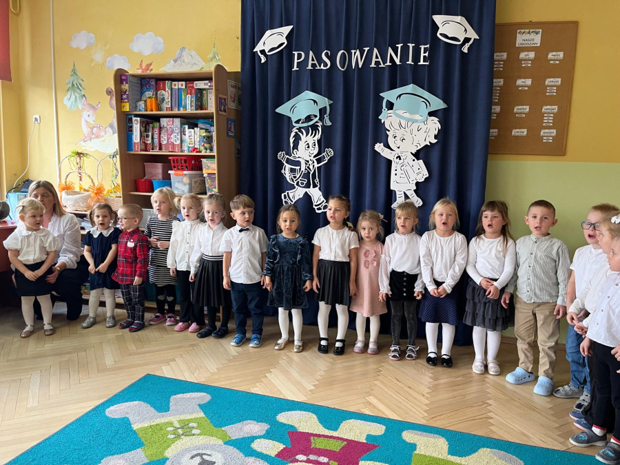 Dzieci stoją w szeregu przy ścianie. Obok nich siedzi nauczycielka a w tle widoczna jest granatowa kurtyna z napisem Pasowanie oraz dekoracje sali