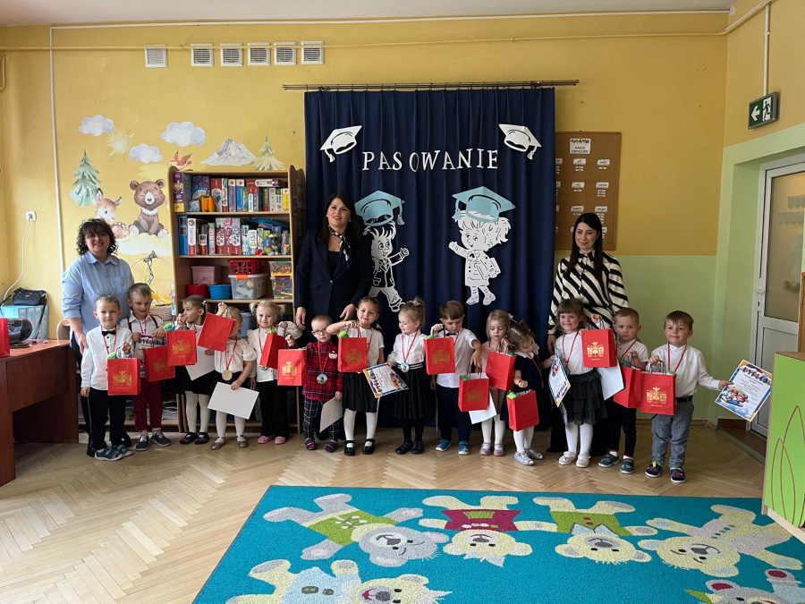 Dzieci stoją w szeregu trzymają w dłoniach czerwone torby i kartki część z nich patrzy w stronę aparatu. Za nimi stoją dorosłe osoby a w tle widoczna jest granatowa kurtyna z dekoracjami