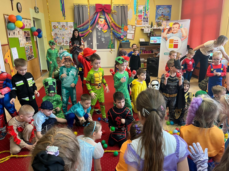 Dzieci w kolorowych przebraniach bawią się na czerwonym dywanie w sali przedszkolnej. Widać stroje superbohaterów zwierząt i postaci z bajek. Na podłodze leżą plastikowe piłeczki