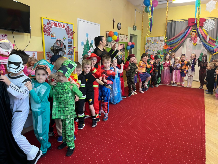 Dzieci w kolorowych przebraniach stoją w rzędzie na czerwonym dywanie w sali przedszkolnej. Wśród strojów widać postacie z bajek superbohaterów i księżniczki. Nauczycielka stoi obok i klaszcze