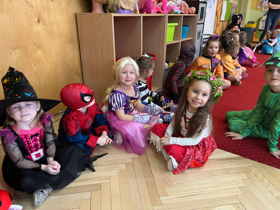 Dzieci w przebraniach siedzą na podłodze przy ścianie w sali przedszkolnej. Widać strój czarownicy Spider Mana księżniczki oraz dziewczynkę w czerwonej sukience z wiankiem na głowie. W tle regały z zabawkami