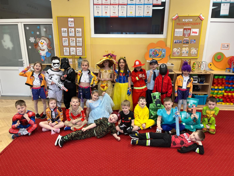 Grupa dzieci w przebraniach stoi i siedzi na czerwonym dywanie. Wśród strojów widać superbohaterów, księżniczki, strażaka i postacie z gier. W tle tablice edukacyjne oraz kącik patriotyczny. 