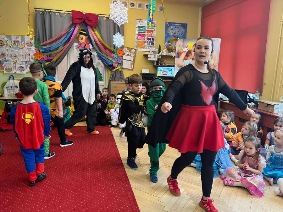 Nauczycielka w stroju z czerwonym emblematem prowadzi dzieci przebrane za superbohaterów i postacie z bajek po czerwonym dywanie. W tle kolorowa dekoracja z kokardą i girlandami a pozostałe dzieci siedzą