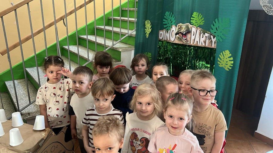 Grupa dzieci stoi obok dekoracji z napisem „Dino Park” i ilustracją dinozaura. Dzieci ustawione są blisko siebie i patrzą w stronę aparatu. Za nimi widoczna jest zielona kurtyna z motywami liści