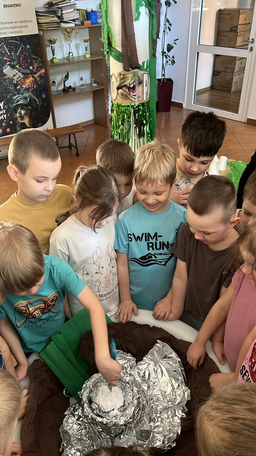 Grupa dzieci stoi wokół okrągłego stolika i obserwuje doświadczenie. Na środku znajduje się model wulkanu wykonany z folii aluminiowej i papieru. Z wnętrza wulkanu wydobywa się biała piana