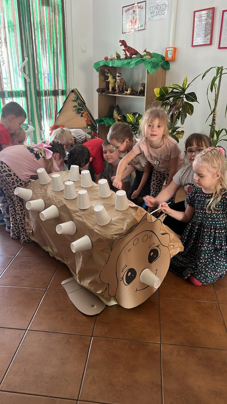 Grupa dzieci zgromadzona wokół dużej kartonowej makiety dinozaura ustawionej na podłodze. Konstrukcja oklejona jest brązowym papierem