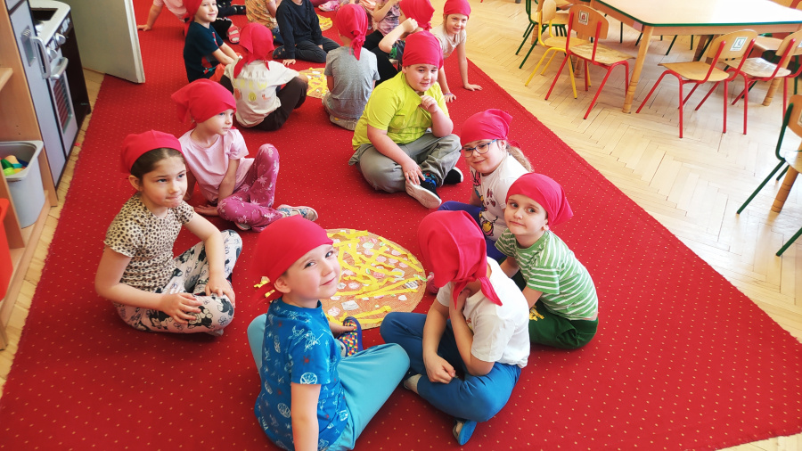 Dzieci siedzą na czerwonym dywanie. Mają na głowach czerwone chustki i pracują nad dużą papierową pizzą ułożoną na środku. W tle stoliki i krzesła 