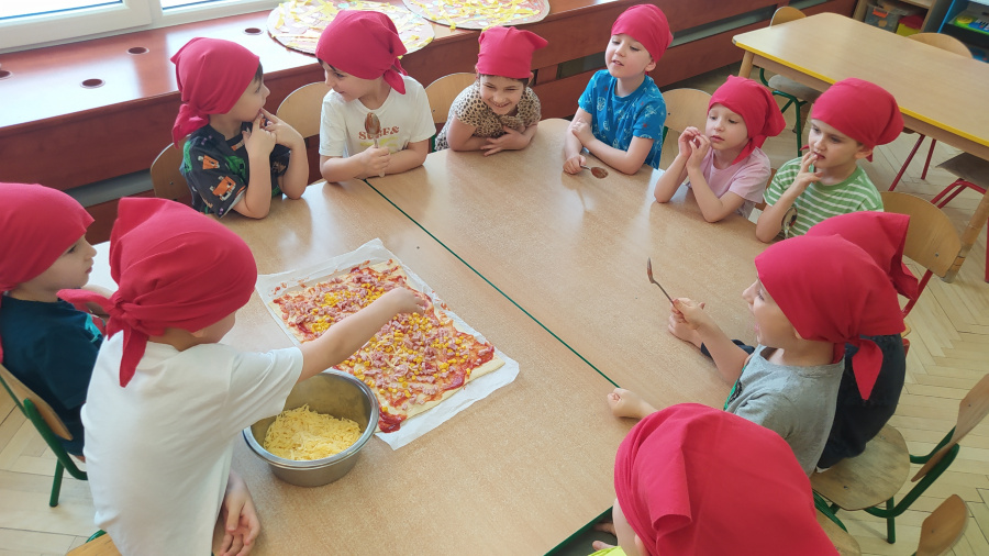Dzieci w czerwonych chustkach siedzi przy stoliku. Na blacie leży duże ciasto z sosem i pokrojonymi dodatkami obok miska z tartym serem. Jedno dziecko posypuje pizzę składnikami pozostałe obserwują