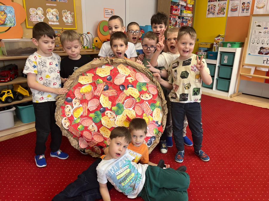 Grupa chłopców stoi na czerwonym dywanie i trzyma dużą okrągłą pracę plastyczną przedstawiającą pizzę z kolorowymi dodatkami. Dwóch chłopców siedzi z przodu pozostali pozują z uśmiechami. W tle półki