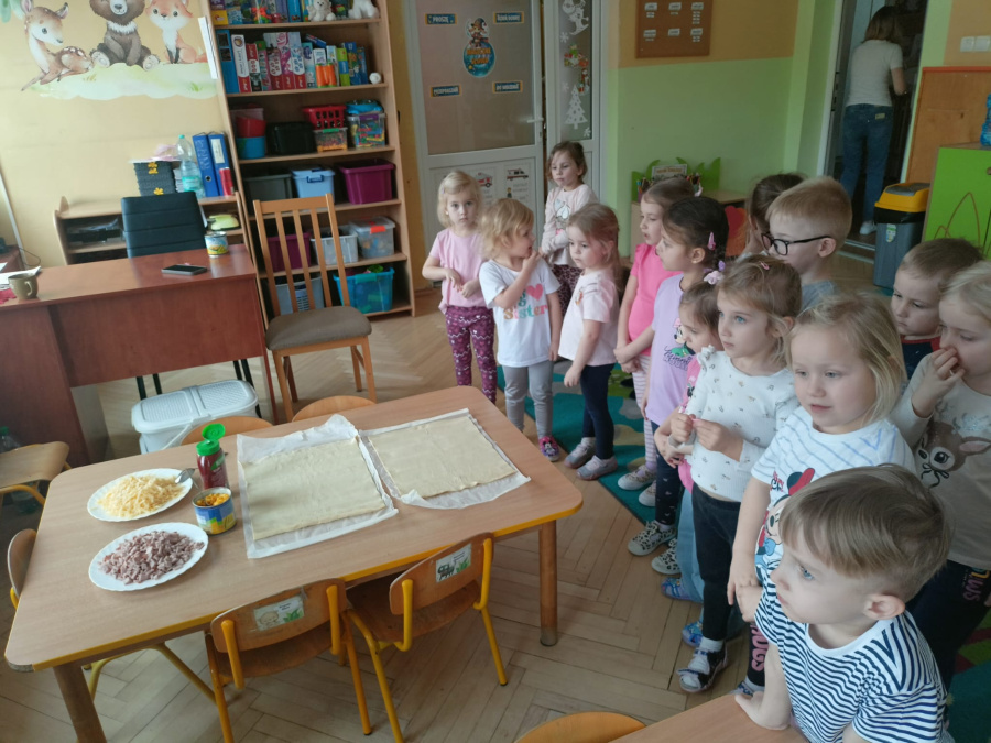 Grupa dzieci stoi przy stoliku. Na blacie leżą dwa prostokątne ciasta talerze z pokrojoną szynką i startym serem oraz butelka sosu. Dzieci z zainteresowaniem obserwują przygotowane składniki. W tle rega