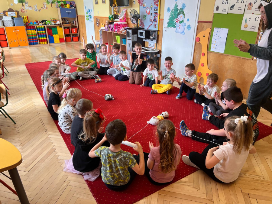 Dzieci siedzą w kręgu na czerwonym dywanie. Każde trzyma drewnianą wędkę ze sznurkiem skierowanym do środka koła gdzie leżą pluszowe zabawki. Część dzieci klaszcze