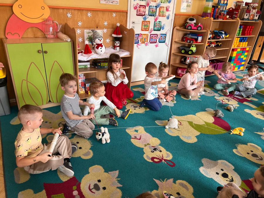 Grupa dzieci siedzi w kręgu na kolorowym dywanie. Każde trzyma drewnianą wędkę z magnesem i łowi pluszowe zabawki przymocowane do sznurków. W tle widoczne półki z zabawkami