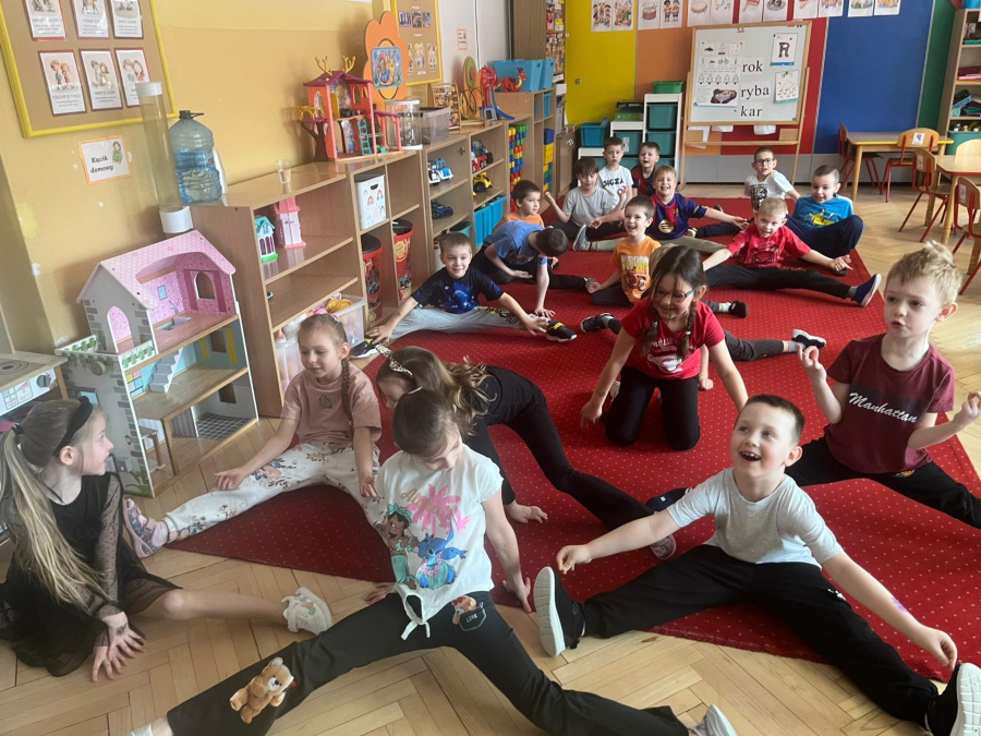 Grupa dzieci ćwiczy na czerwonym dywanie. Większość siedzi w szerokim rozkroku wykonując skłony i rozciąganie. Dzieci uśmiechnięte i zaangażowane w aktywność ruchową