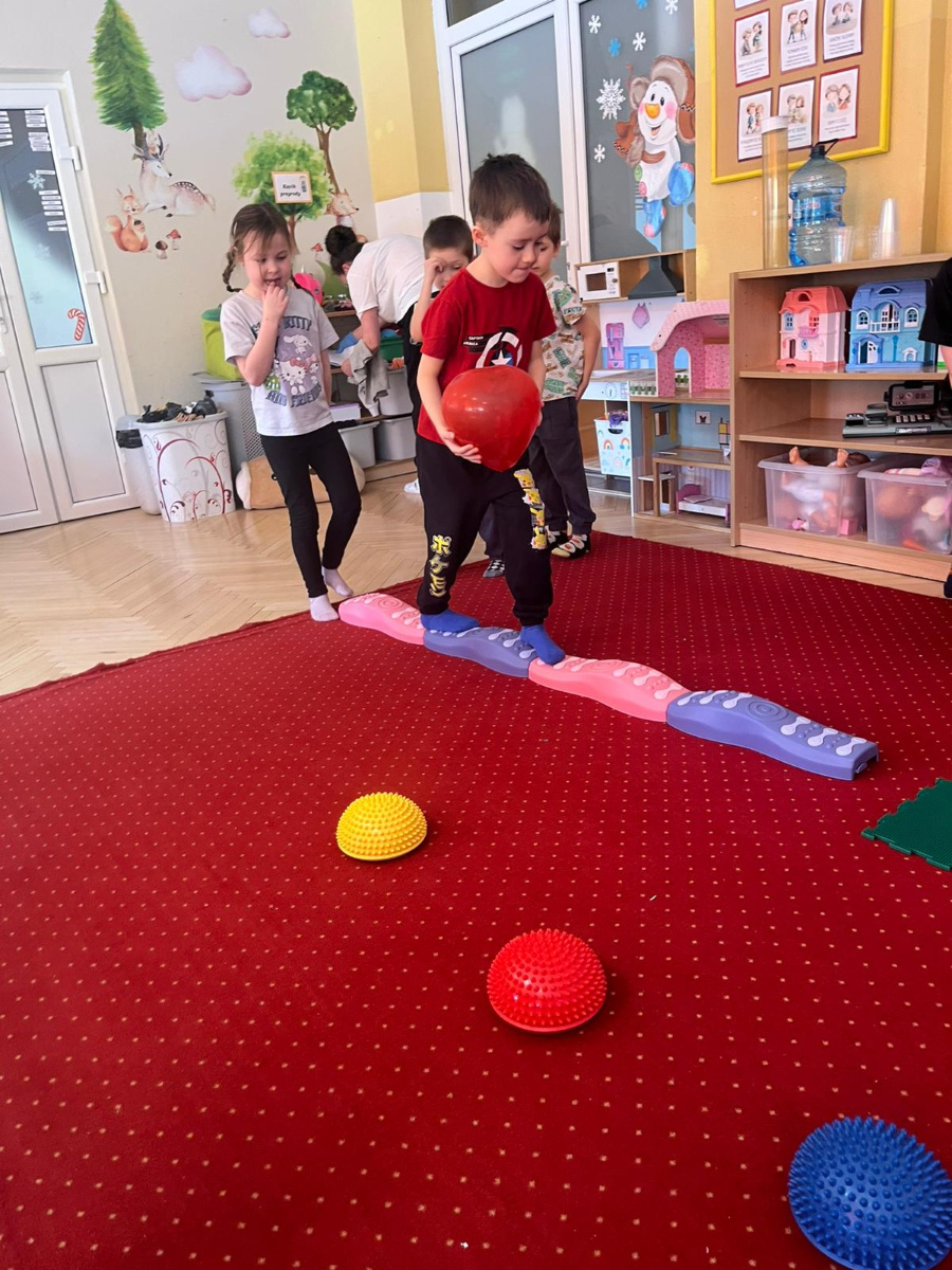 Chłopiec w czerwonej koszulce przechodzi po kolorowej ścieżce sensorycznej trzymając w rękach czerwony balon. Za nim czekają kolejne dzieci obserwując zadani