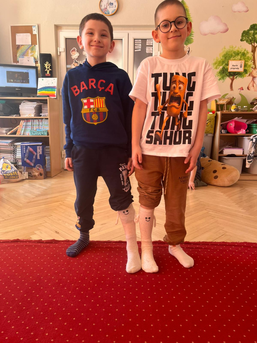 Dwóch chłopców stoi obok siebie na czerwonym dywanie i uśmiecha się do zdjęcia. Jeden ma granatową bluzę drugi białą koszulkę z kolorową grafiką i brązowe spodnie. Obaj mają na nogach skarpetki a w tle w