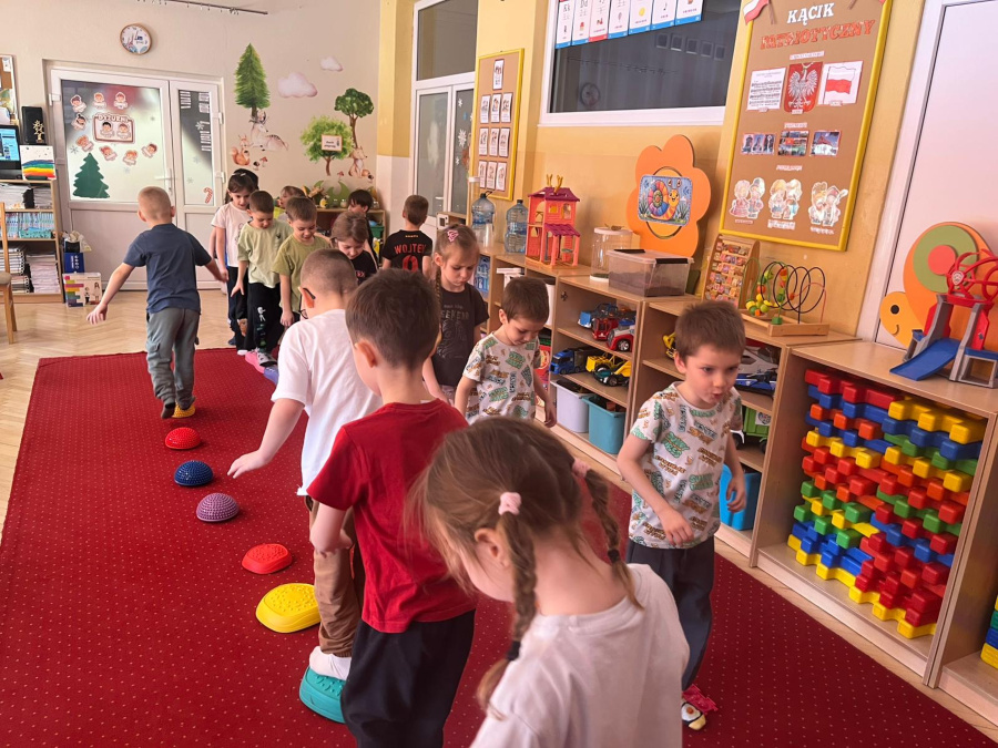 Grupa dzieci idzie po czerwonym dywanie, stawiając stopy na kolorowych półkulach sensorycznych ułożonych w linii. W tle sala przedszkolna z półkami pełnymi zabawek, klocków 