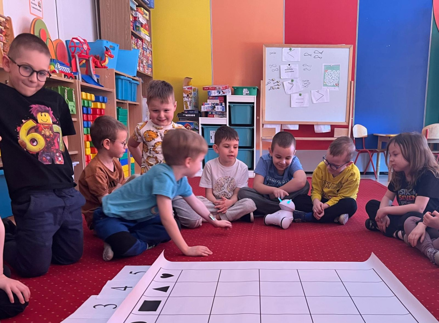 Grupa dzieci siedzi na czerwonym dywanie wokół małego białego robota. Jedno z dzieci dotyka robota a pozostałe obserwują jego działanie. Na podłodze leży duża mata podzielona na kratki W tle znajdują s